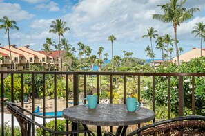 A balcony with a view of a pool and palm trees, a table with two chairs, and a railing. - South Kihei condo with partial ocean view, pool, hot tub, and lanai (Kihei)