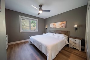 A bedroom with a bed, a nightstand, a ceiling fan, a window, and a painting on the wall.