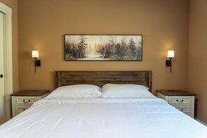 Une chambre à coucher avec une tête de lit en bois, deux tables de chevet, un couvre-lit blanc et un tableau représentant un paysage forestier accroché au mur.