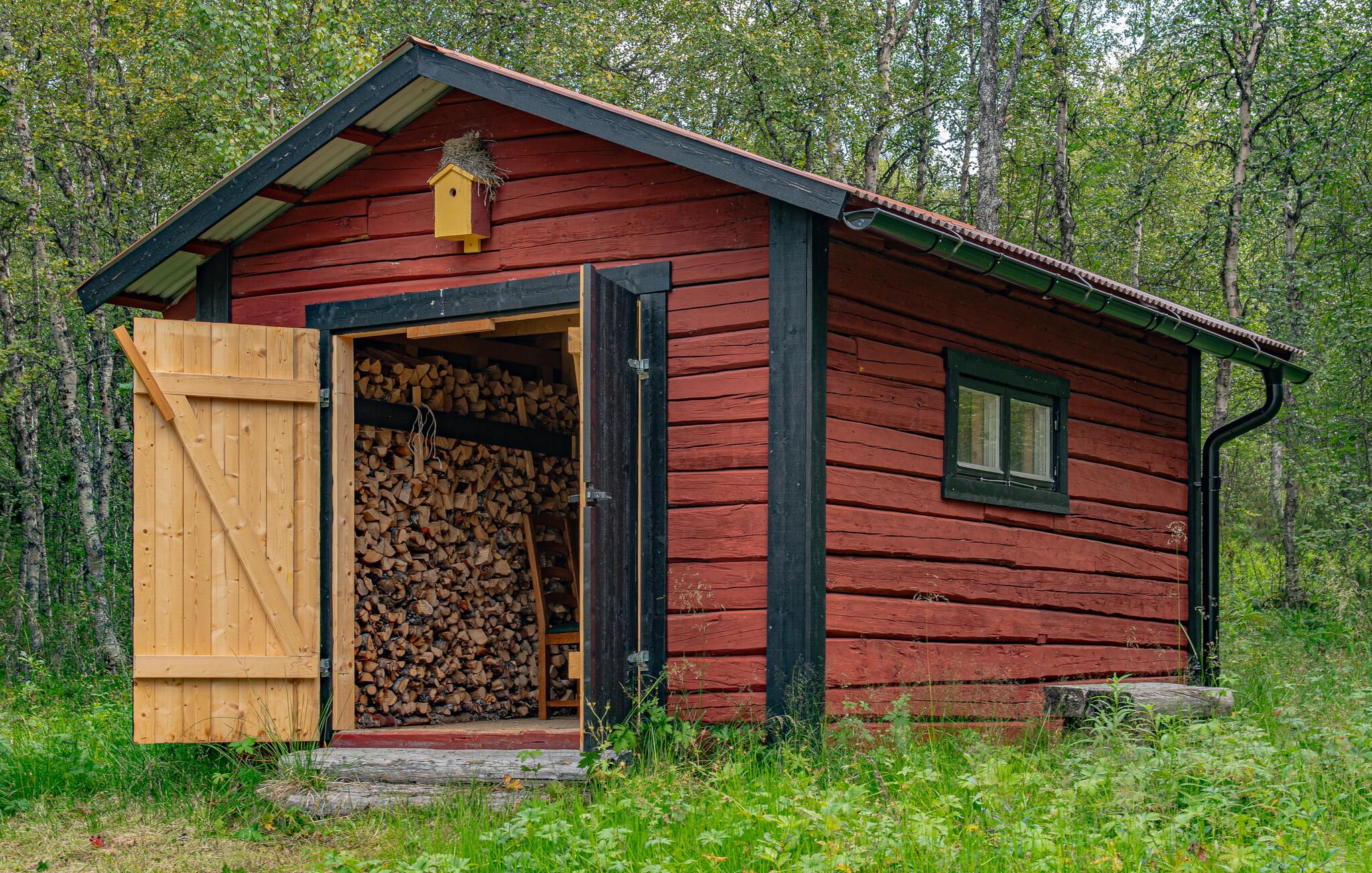 Une petite cabane en bois, dont la porte est ouverte et laisse apparaître une pile de bois de chauffage.