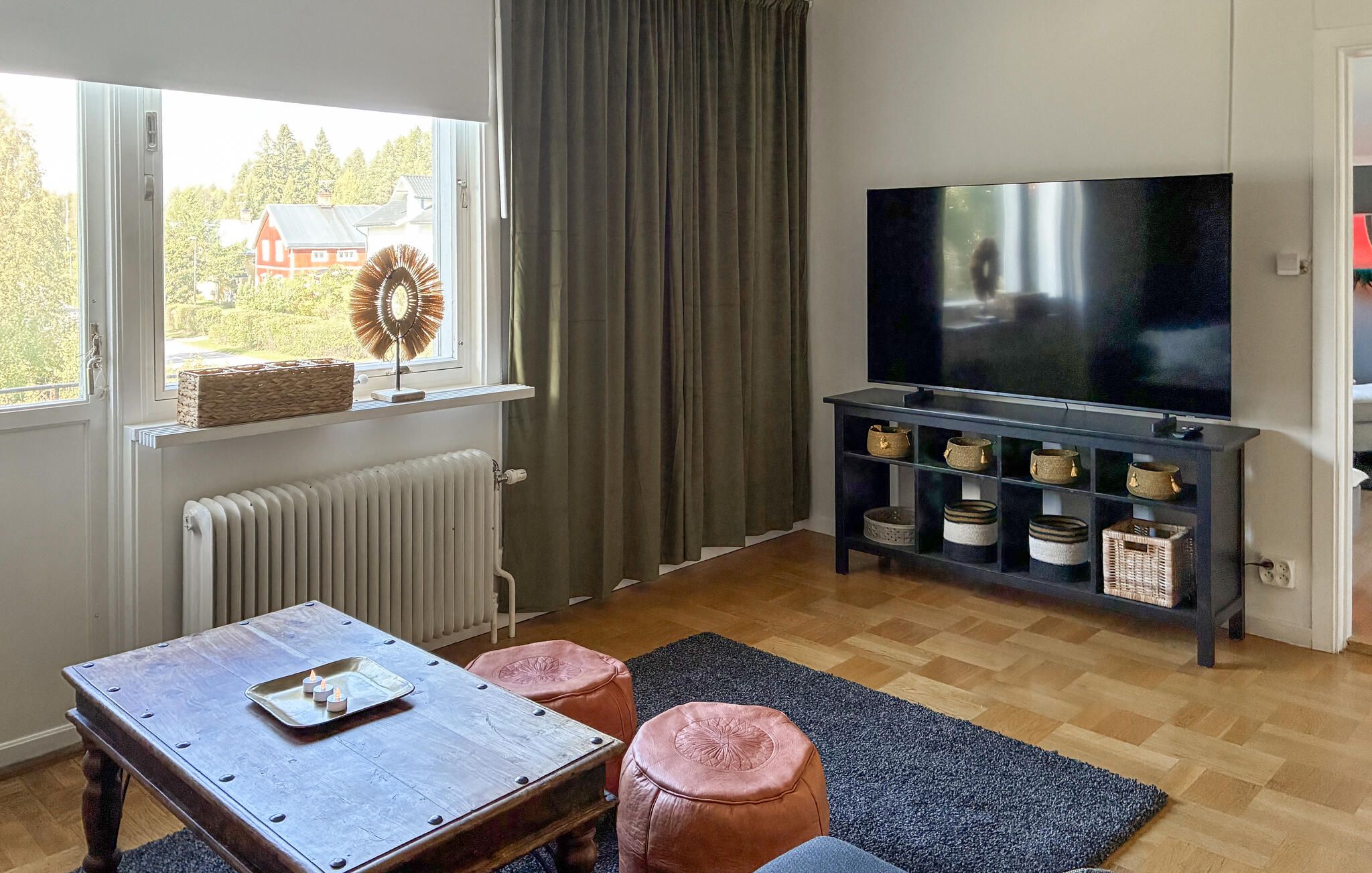 A living room with a wooden table, two leather ottomans, a TV on a stand, and a window with a view of houses.