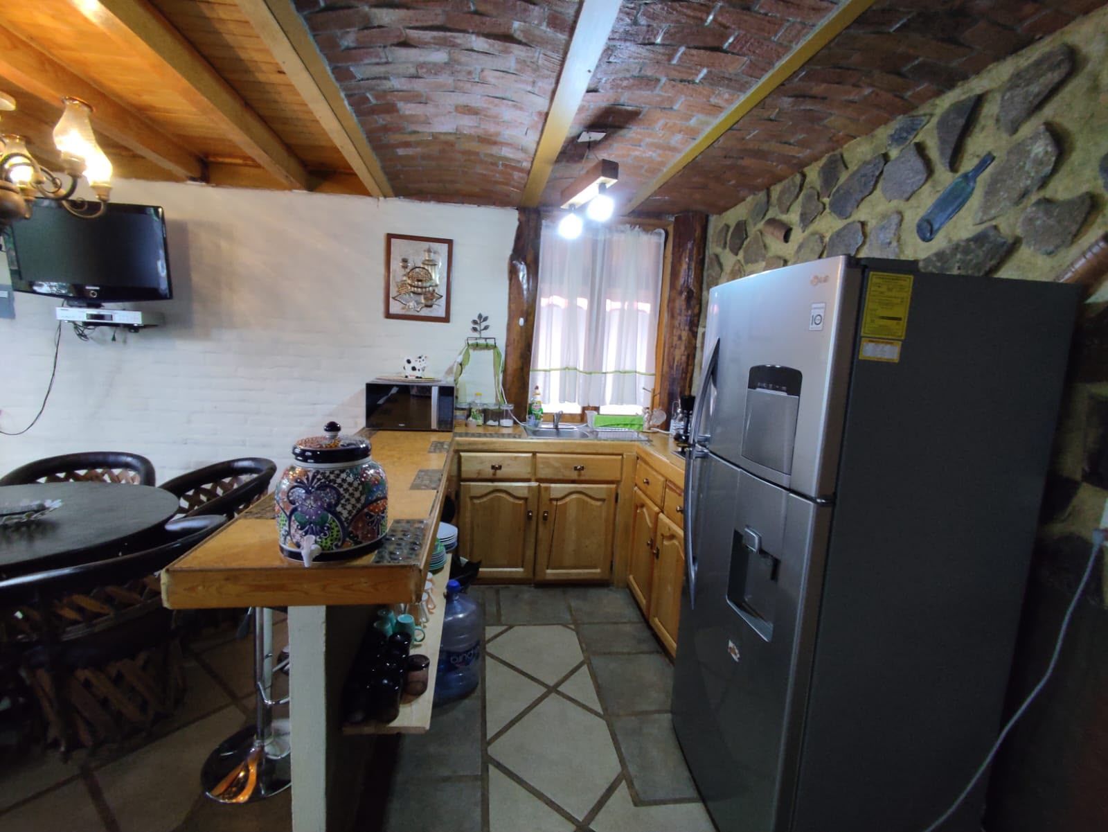 Een keuken met een stenen muur, houten keukenkastjes, een koelkast en een eettafel met stoelen.