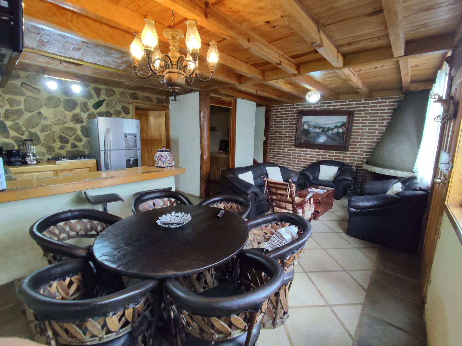Een landelijke inrichting met een stenen muur, houten plafond, een eethoek met een ronde tafel en stoelen, een keuken met een koelkast en een open haard.