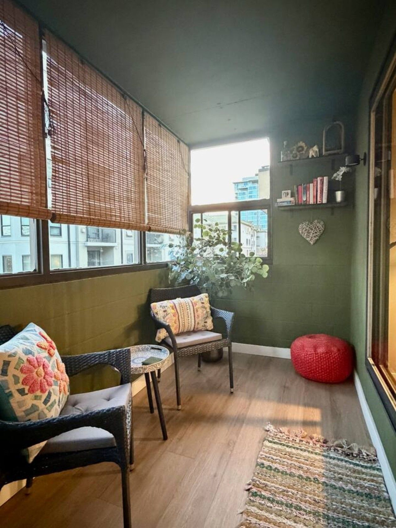 A cozy room with a large window, a wicker chair, a small table, a red bean bag, and a green wall with a heart-shaped decoration.