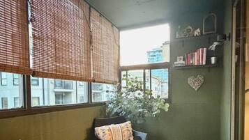 A cozy room with a large window, a wicker chair, a small table, a red bean bag, and a green wall with a heart-shaped decoration.