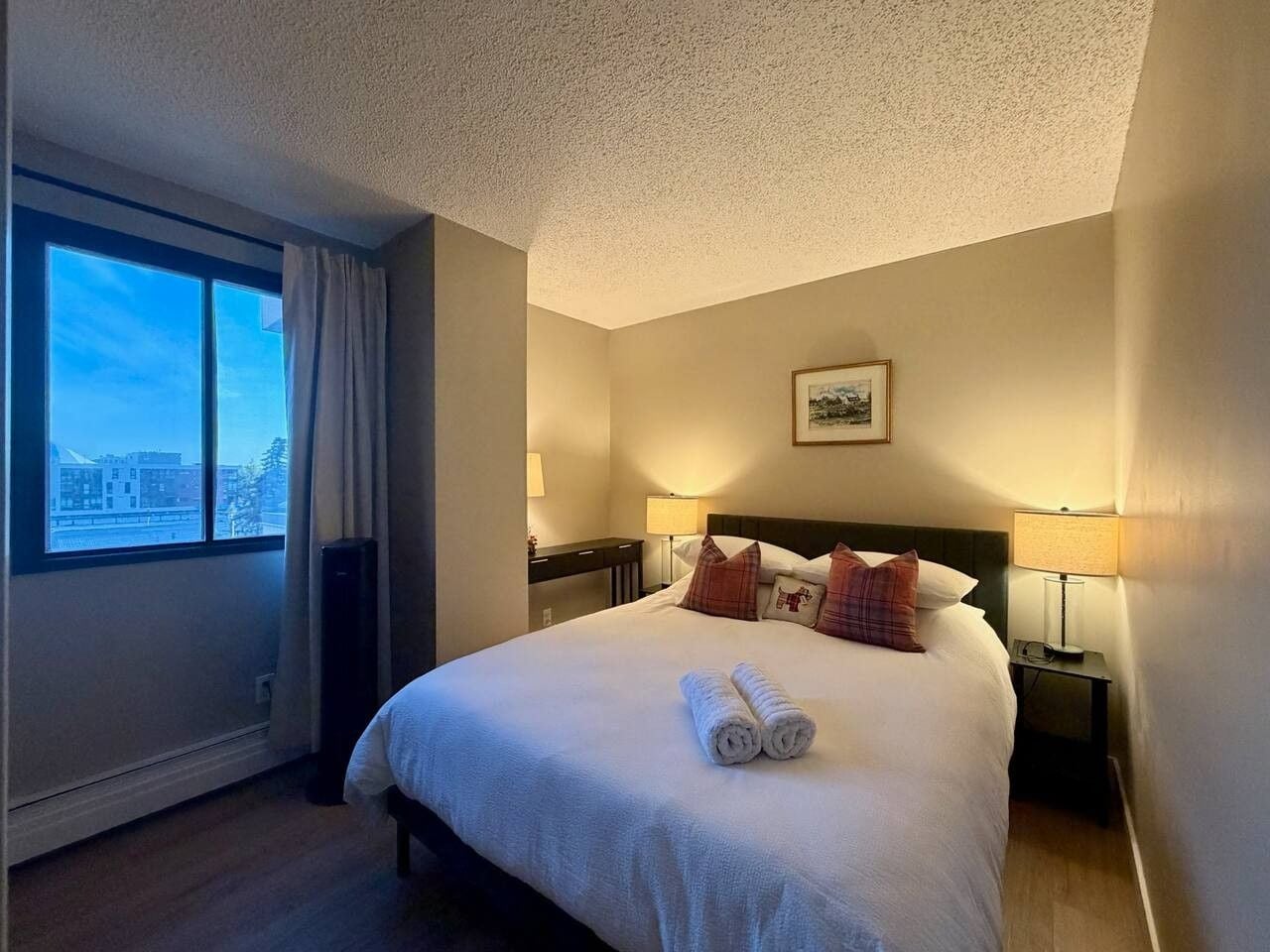 A hotel room with a large bed, two bedside lamps, a window with curtains, and a framed picture on the wall.