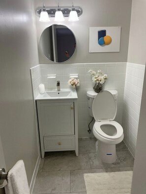 Ein kleines Badezimmer mit Waschbecken, Waschtisch und Toilette, einem runden Spiegel über dem Waschbecken und einer kleinen dekorativen Vase mit Blumen.