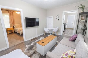 A modern living room with a flat-screen TV, a sofa, a coffee table, and a dining area with chairs. - Porchlight Cottage - South Bend, IN (South Bend)