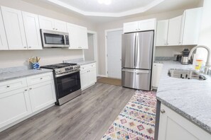 A modern kitchen with white cabinets, a stainless steel refrigerator, a microwave, an oven, and a granite countertop. - Porchlight Cottage - South Bend, IN (South Bend)