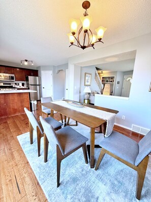 A modern kitchen with stainless steel appliances, a dining area with a wooden table and grey upholstered chairs, and a chandelier with multiple light fixtures. - 3 Bedroom Family Home in NW Calgary (Calgary)