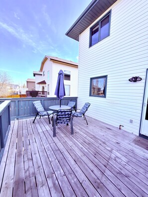 A wooden deck with a table and chairs, a covered umbrella, and a house with white siding and windows. - 3 Bedroom Family Home in NW Calgary (Calgary)