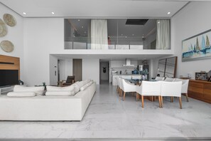 Uma sala de estar moderna com um sofá grande, área de jantar com cadeiras brancas e uma cozinha ao fundo.
