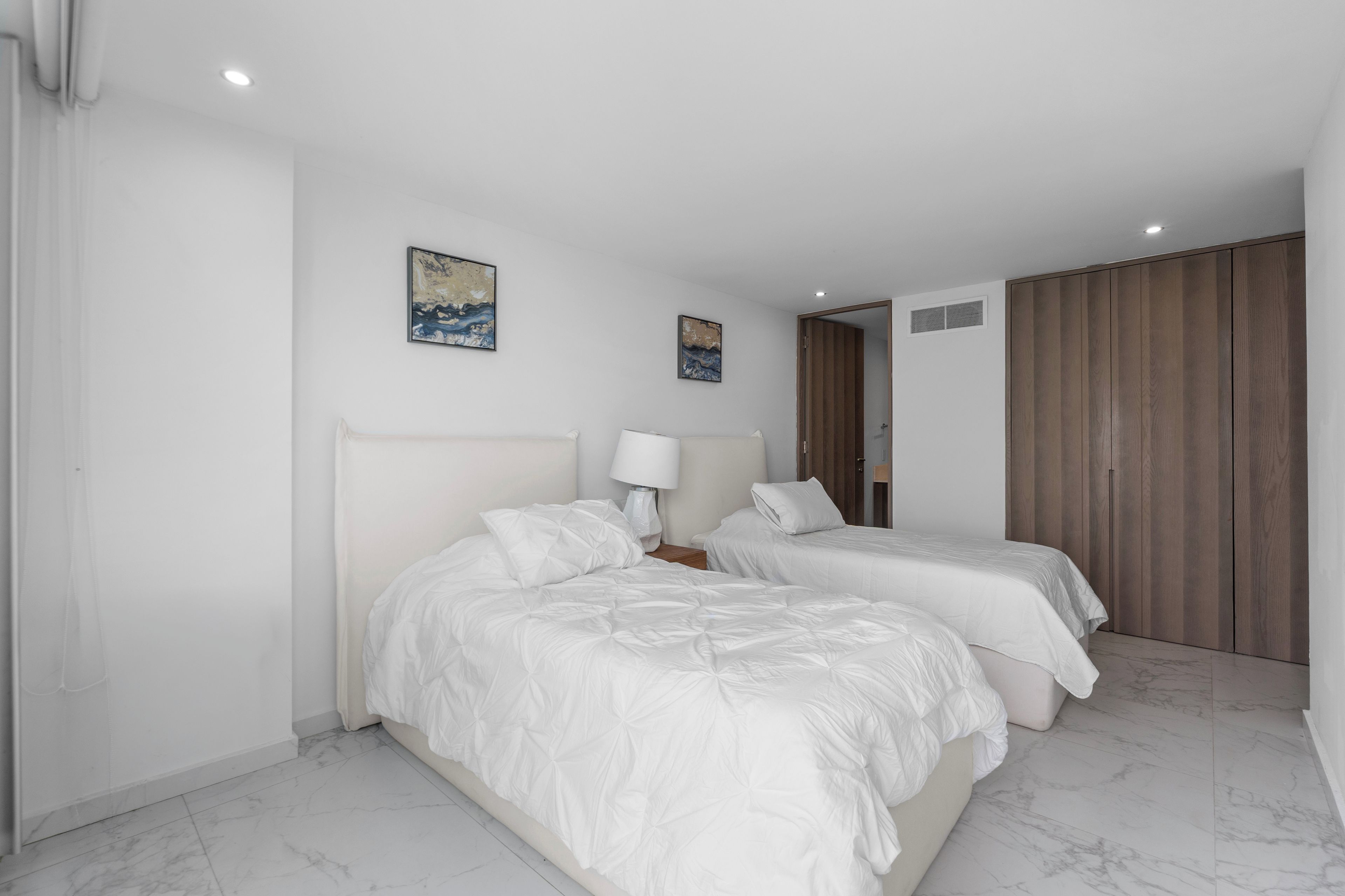 A hotel room with two beds, a nightstand, a lamp, and two paintings on the wall.