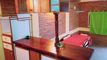 Una habitación compacta con un escritorio de madera, una mesita con mantel rojo, una botella verde y una pared de ladrillo.