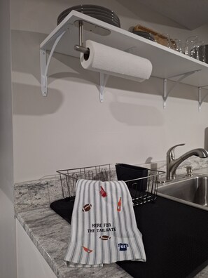 Une cuisine dotée d’un plan de travail en marbre, d’un évier en acier inoxydable, d’un porte-serviettes avec une serviette à rayures et d’une étagère où sont rangées des assiettes et des verres.