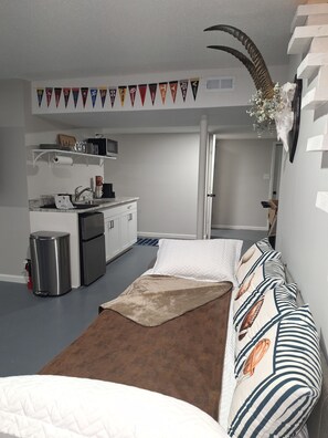 Una habitación pequeña con kitchenette, una cama con almohadones estampados y un adorno en la pared.