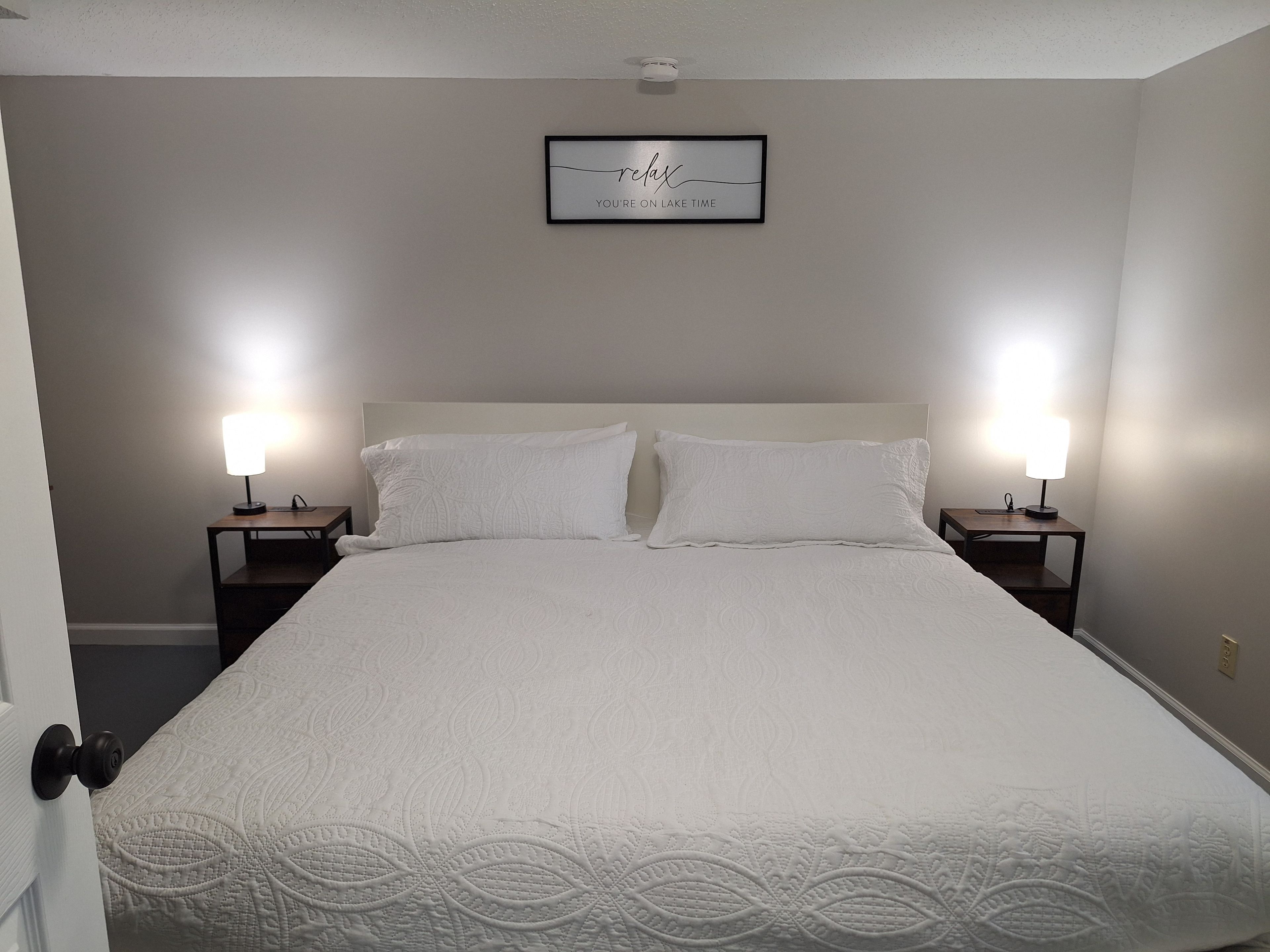 Ein ordentlich bezogenes Bett mit weißer Decke und zwei Kissen, seitlich flankiert von Nachttischen mit Lampen, in einem Zimmer mit schlichter Wand und einem gerahmten Schild darüber.