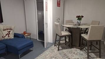 A room with a blue sofa, a dining table with chairs, a patterned rug, and a white wardrobe with a red emergency exit sign.