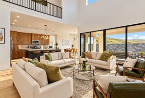 A spacious living area with a high ceiling, a kitchen in the background, and a large window offering a view of the outdoors. - Hurricane Hideaway - Hot Tub + Ping Pong - Pet Ok! (Hurricane)