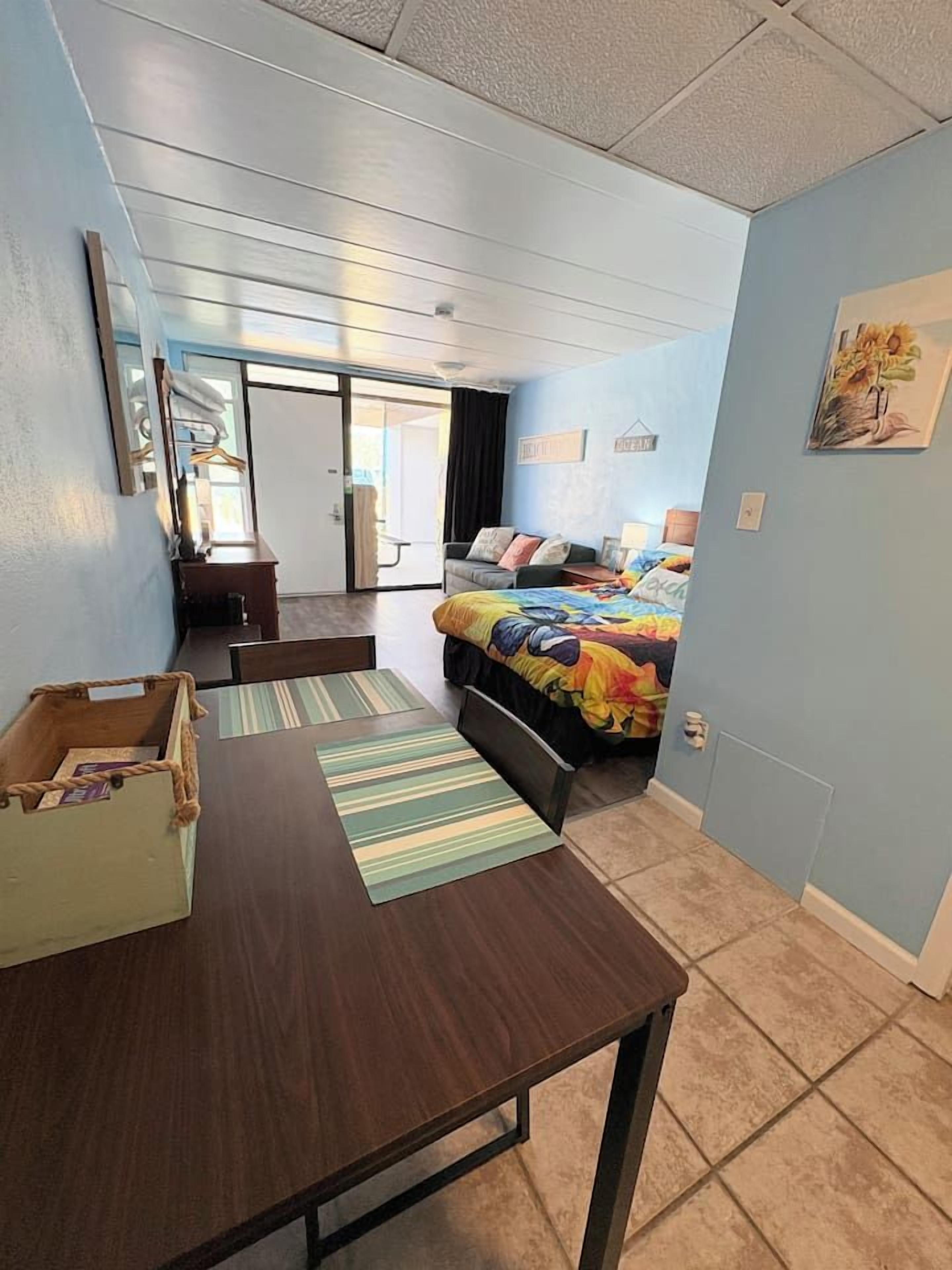 A room with a bed, a desk, and a chair. There is a door leading to another room.