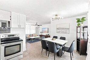 Eine moderne Küche mit eingebauten Geräten, ein Essbereich mit einem weißen Tisch und schwarzen Stühlen sowie ein Wohnbereich mit einer Couch und einem Deckenventilator.