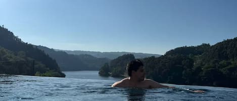 Una persona nada en una piscina azul cristalina, con vista a una ladera boscosa y una extensión de agua en la distancia.
