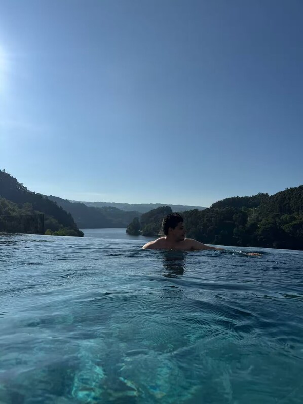 A person swimming in a clear blue pool with a view of a forested hillside and a body of water in the distance.