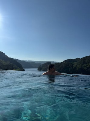 A person swimming in a clear blue pool with a view of a forested hillside and a body of water in the distance.