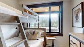 A room with a bunk bed, a small table, and a window with a view of trees. - Switchback Creek Two #5 | 3 Peaks Rentals (Sun Peaks)