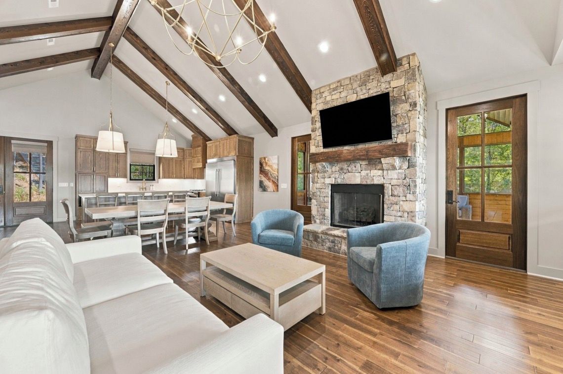 A spacious living area with a stone fireplace, wooden beams, and a dining area with a kitchen in the background.