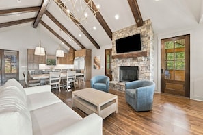 A spacious living area with a stone fireplace, wooden beams, and a dining area with a kitchen in the background.
