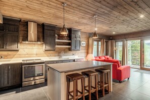 A modern kitchen with a central island, bar stools, and a red sofa in a bright room with wooden ceilings and large windows. - Private Luxury Lodge on Lac Sacacomie, (Saint-Alexis-des-Monts)