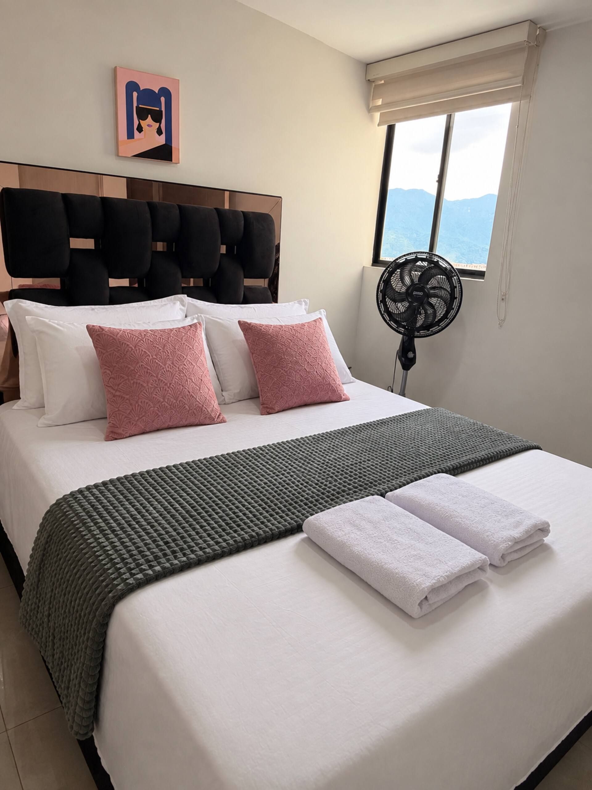 Een hotelkamer met een bed, kussens, dekens, een ventilator en een raam met uitzicht.