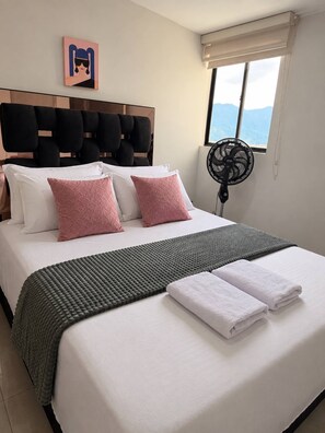 Een hotelkamer met een bed, kussens, dekens, een ventilator en een raam met uitzicht.