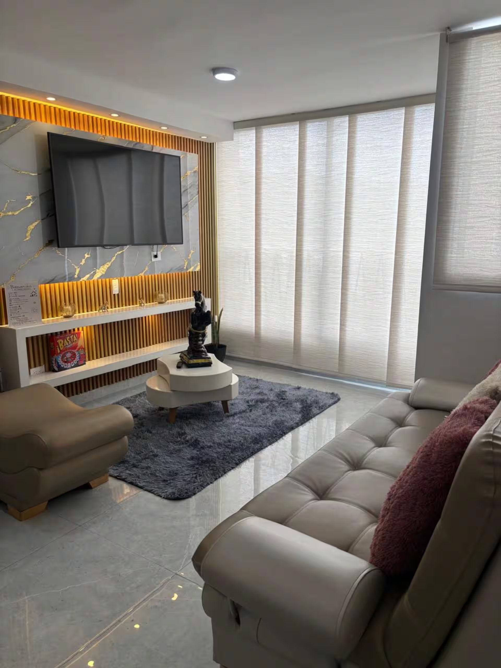 Een moderne woonkamer met een flatscreen televisie aan een wand met een marmerachtig patroon, een bank, een salontafel en een groot raam met gordijnen.