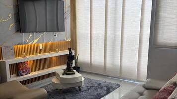 Een moderne woonkamer met een flatscreen televisie aan een wand met een marmerachtig patroon, een bank, een salontafel en een groot raam met gordijnen.