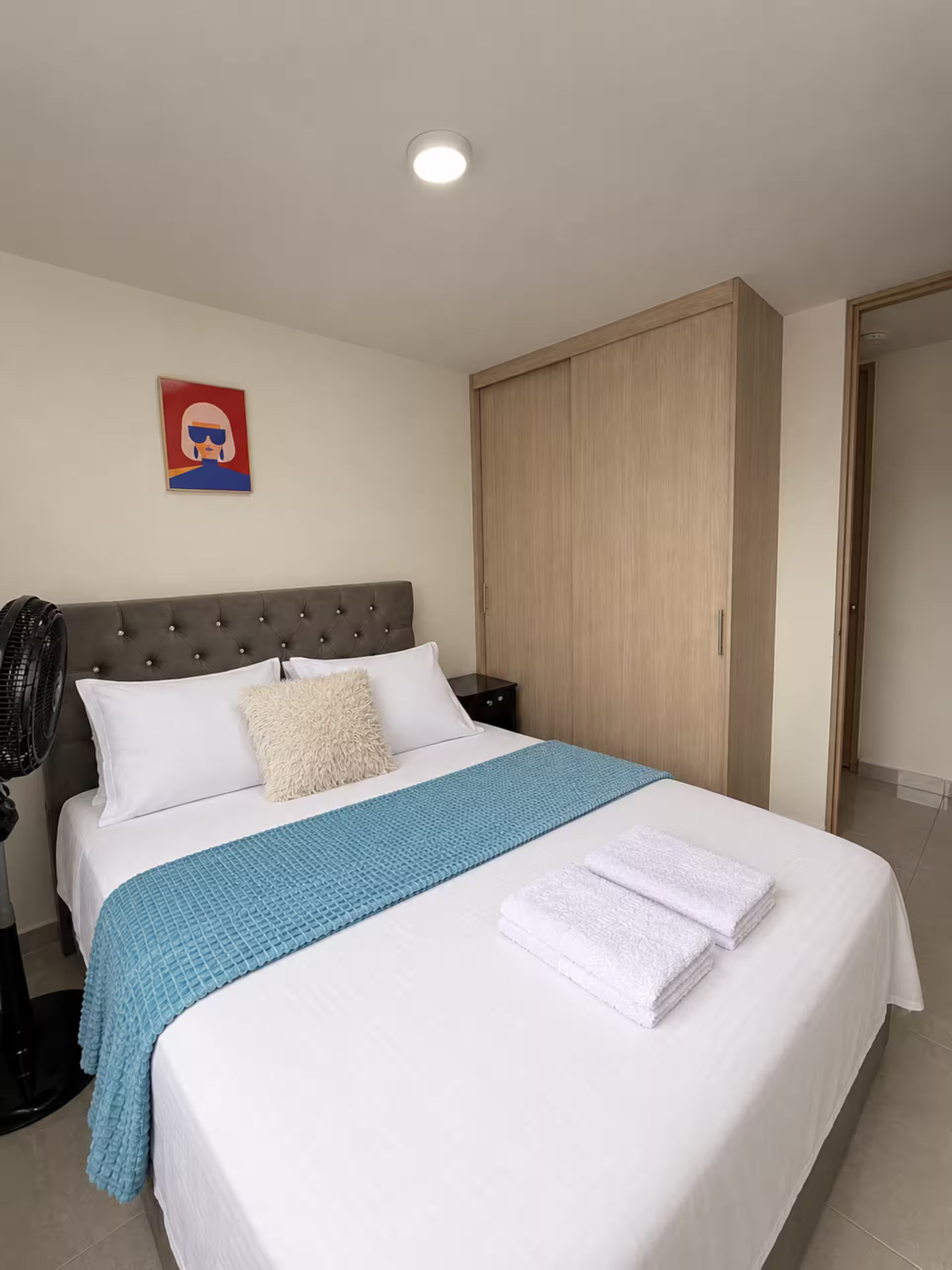 Habitación de hotel con cama, mesita de noche, ventilador de pared, armario y luz de techo.