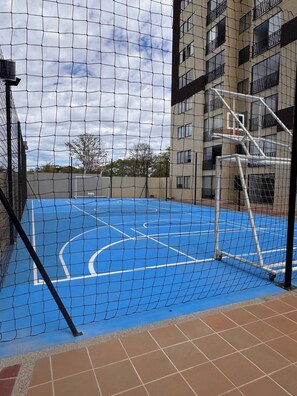 Een buitenbasketbalveld met een net, omringd door een hek, grenzend aan een meerhoog gebouw met balkons.