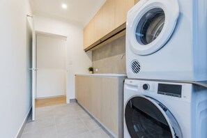 A laundry room with a washing machine and dryer, wooden cabinets, and a tiled floor. - Coastal Haven | Boat Parking | Walk to Town (Agnes Water)