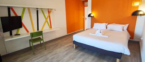 A hotel room with a bed, a desk with a television, a chair, and a wall with colorful lines.