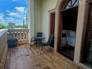 A balcony with a wooden floor, a small table with chairs, a potted plant, and a view of greenery and buildings. - Hibisco Hotel Boutique (Encarnación)