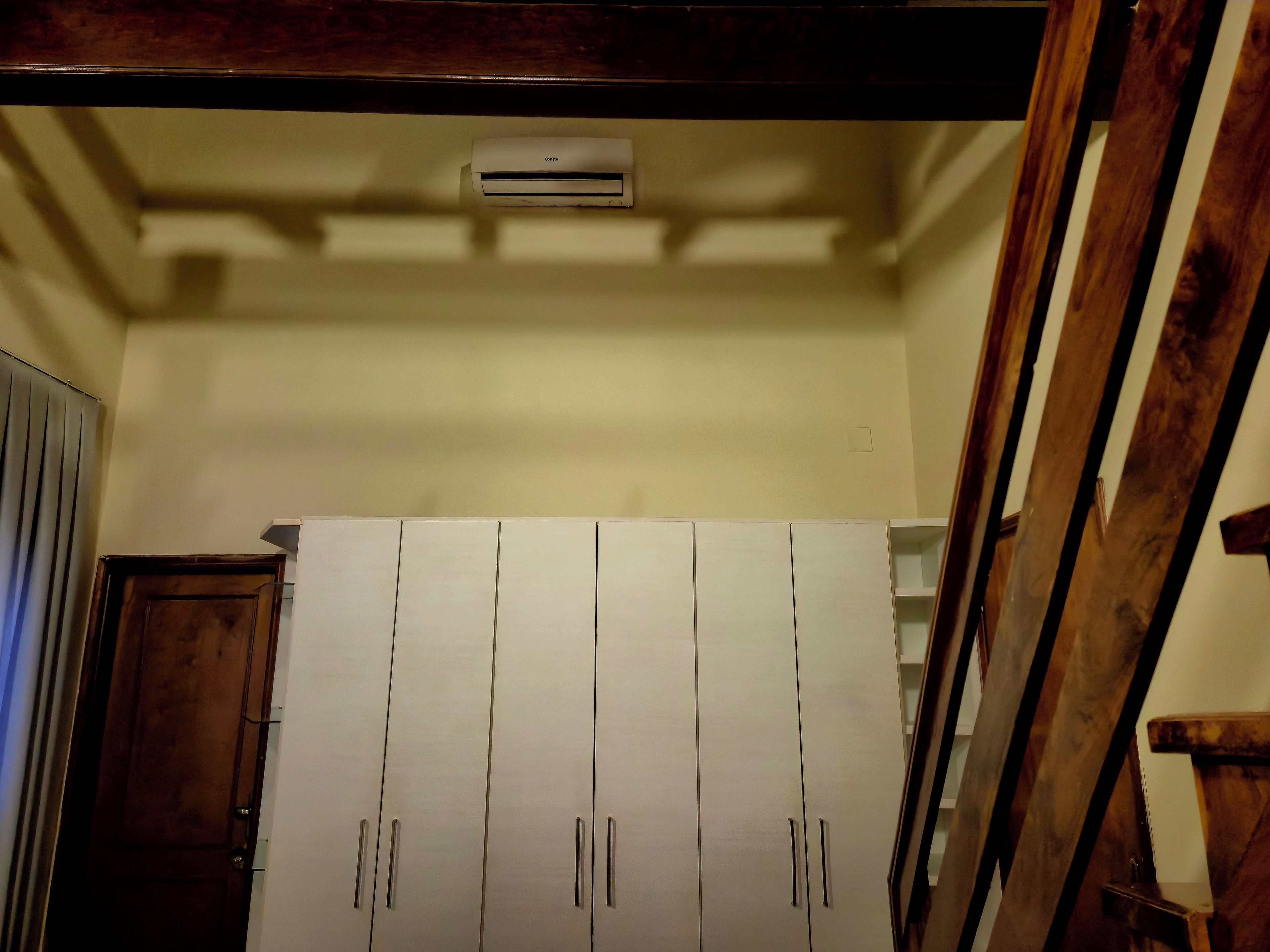 A room with a white wardrobe, a wooden door, and a ceiling-mounted air conditioning unit.