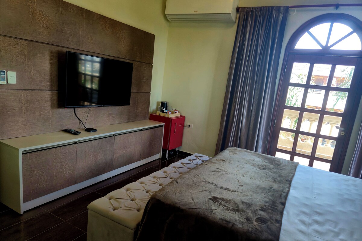A hotel room with a bed, a TV mounted on a wooden panel, a red cabinet, and a window with curtains.