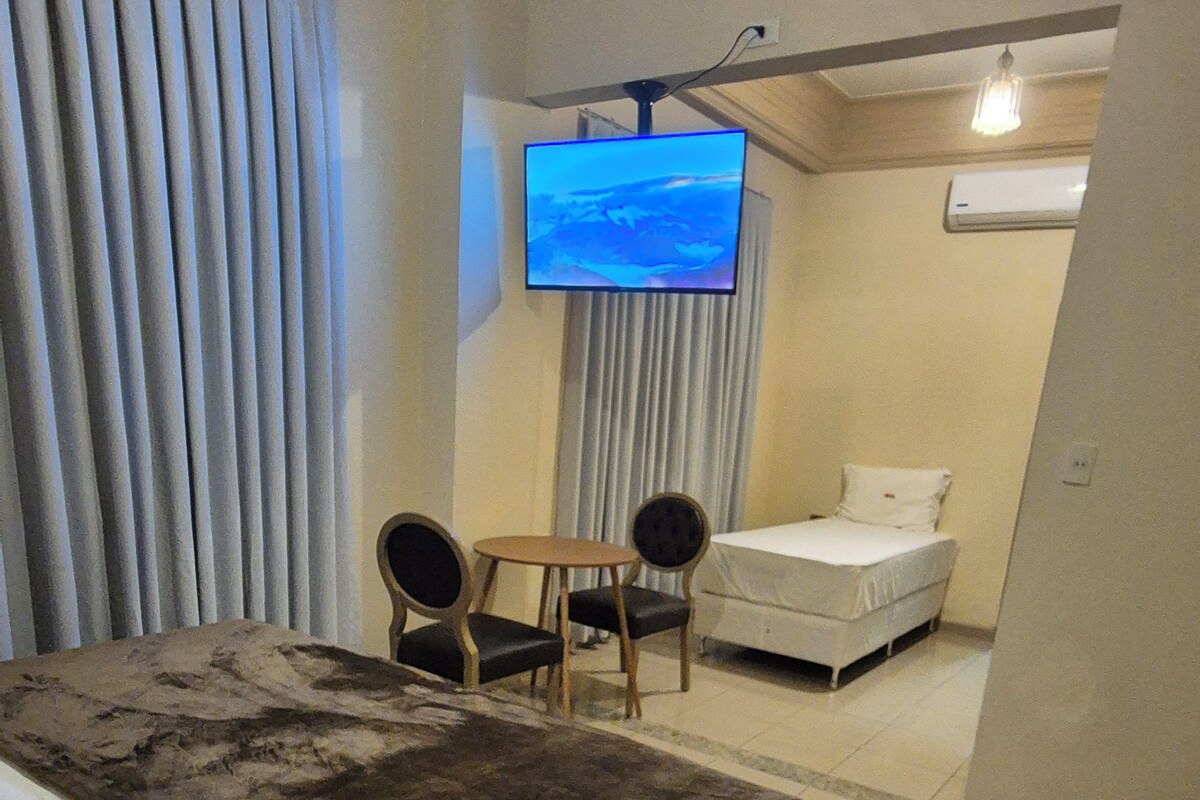 A hotel room with a bed, a TV mounted on the wall, a small table with chairs, and a separate sleeping area with a bed.