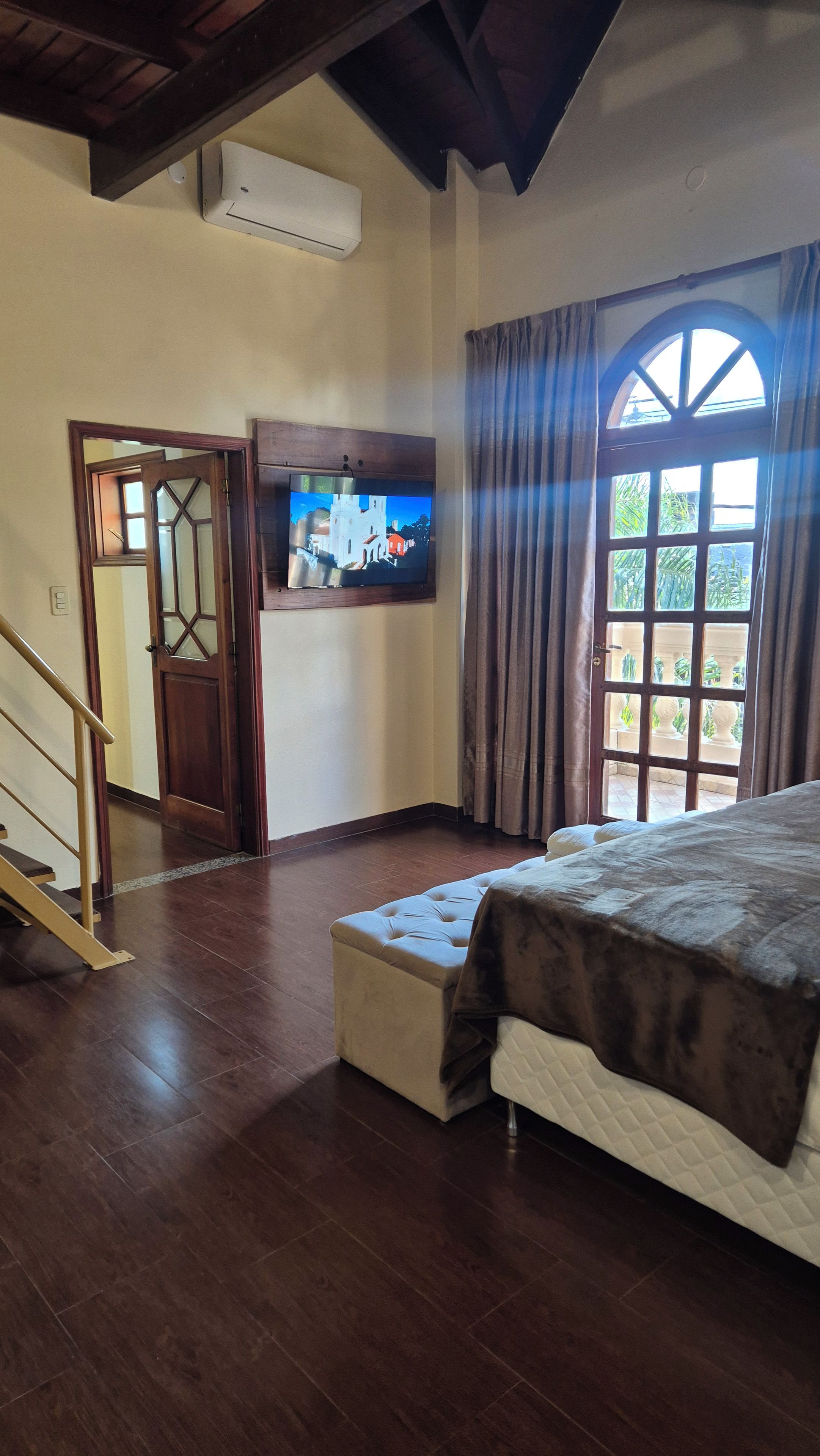 A bedroom with a bed, a television, a window with curtains, and a staircase.