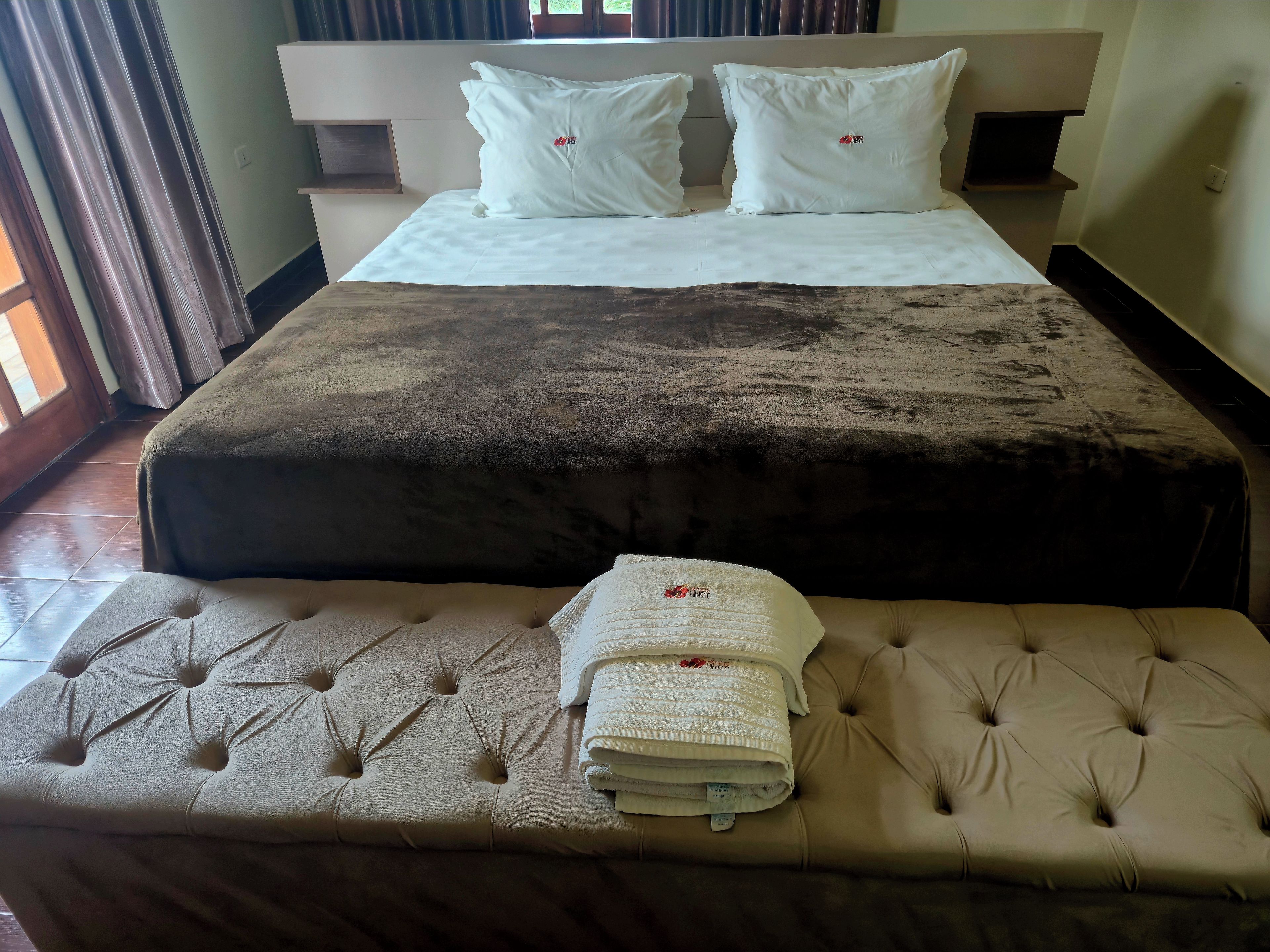Una cama bien hecha con una colcha marrón y dos almohadas blancas, una mesita de noche con una toalla blanca doblada, y una ventana con cortinas.