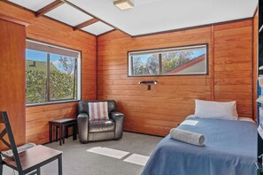 Une pièce aux murs en bois, un lit avec une literie blanche, une chaise avec un coussin à rayures et une fenêtre donnant sur des arbres.