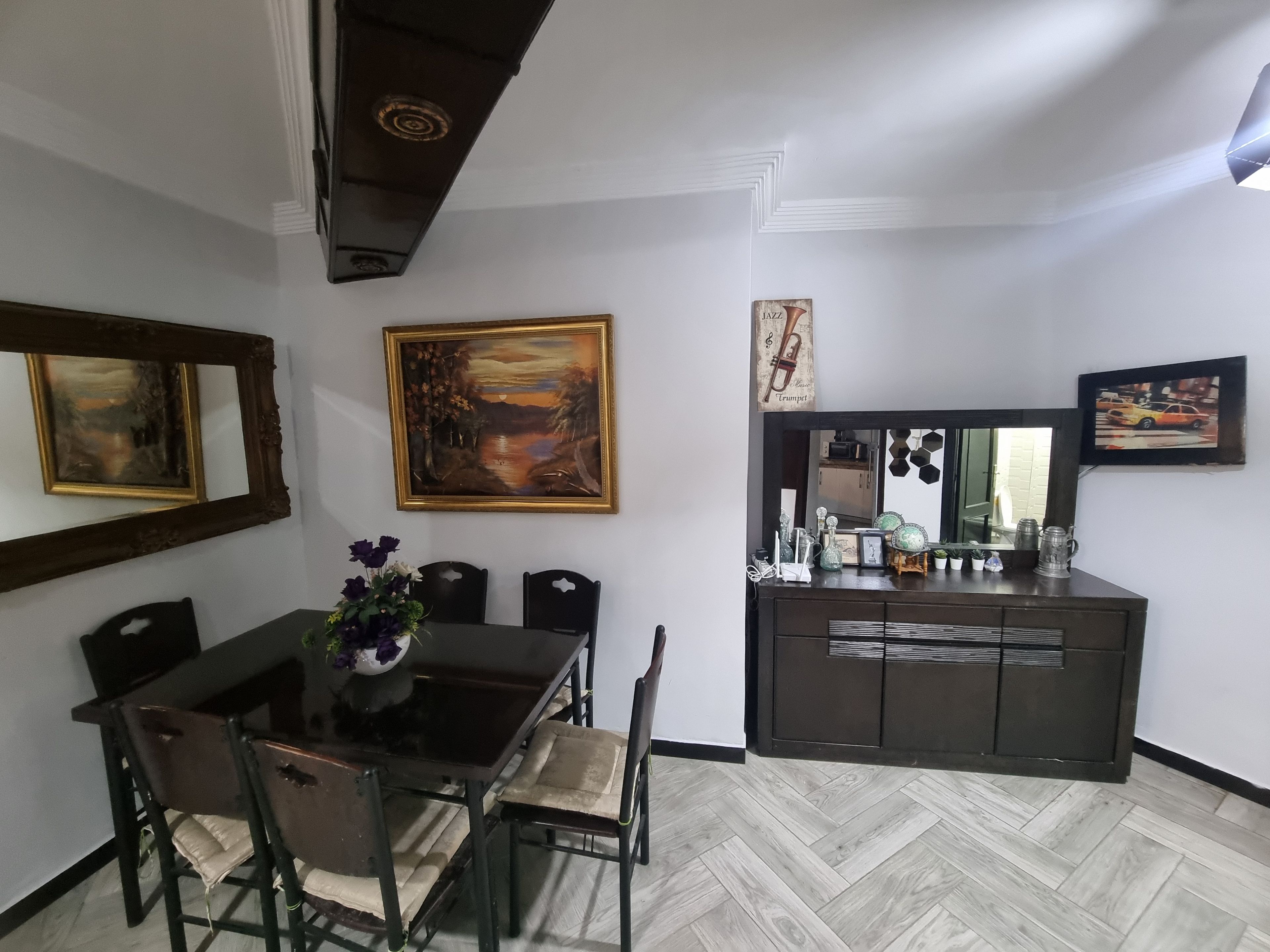 A dining area with a table and chairs, a painting, a mirror, and a cabinet with decorative items.