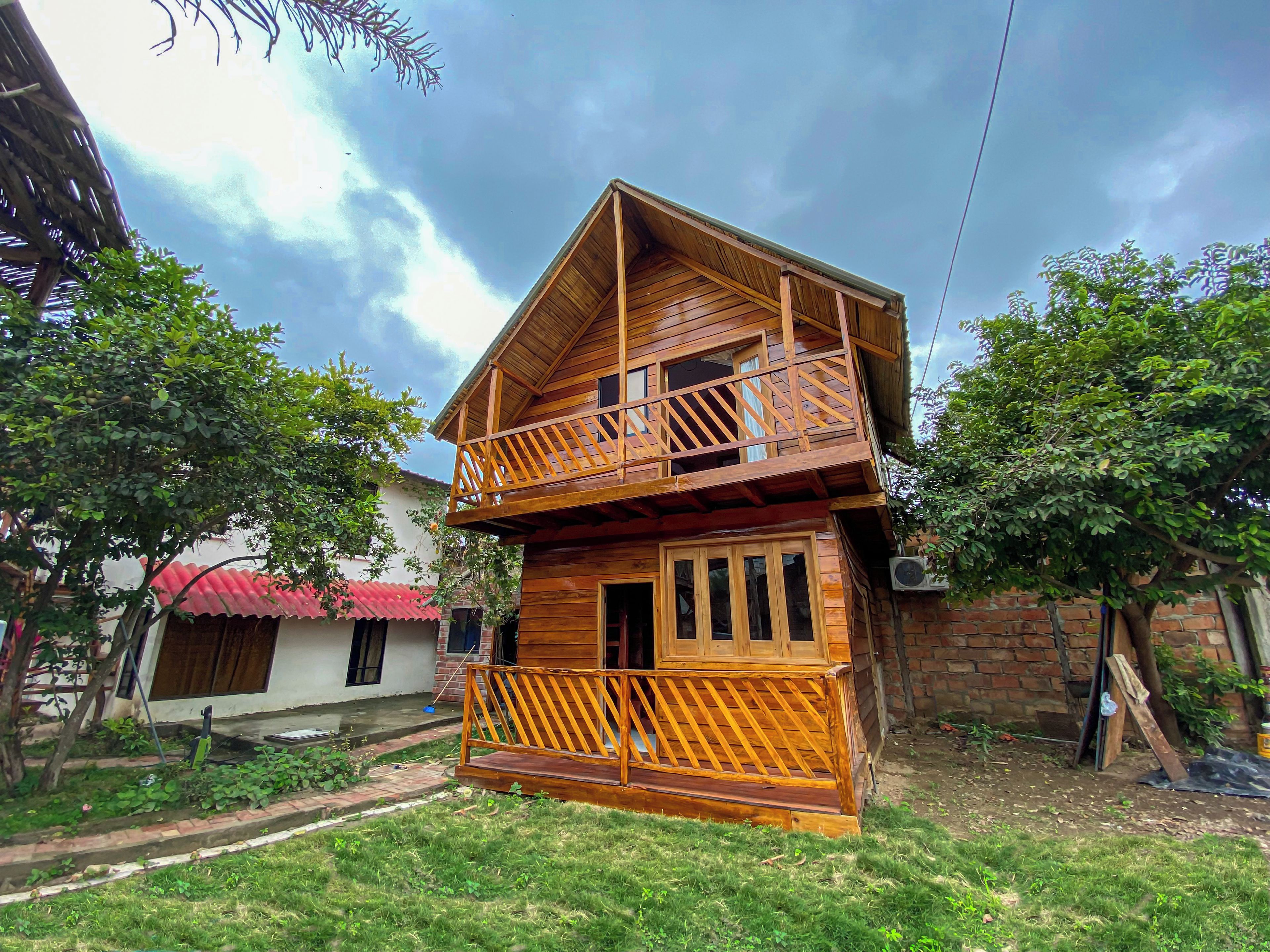 A two-story wooden house with a balcony, surrounded by greenery and a cloudy sky.