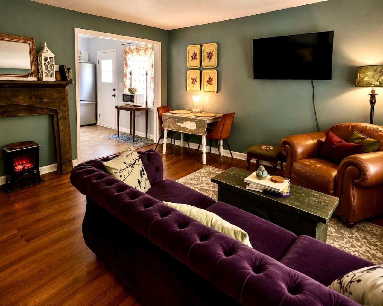 Ein Wohnzimmer mit violettem Sofa, Kamin, Essbereich mit Tisch und Stühlen sowie einem an der Wand befestigten Fernseher.